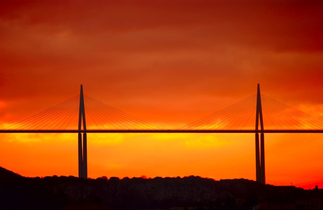 Viaduto de Millau elevando-se acima da paisagem montanhosa francesa, evidenciando sua estrutura estaiada, os pilares monumentais e a integração visual com o ambiente natural ao redor