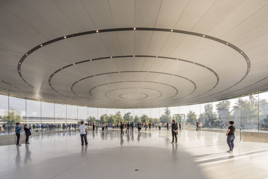Salão circular do Apple Park, com fachada de vidro do piso ao teto, cobertura com anéis concêntricos e visitantes distribuídos pelo espaço
