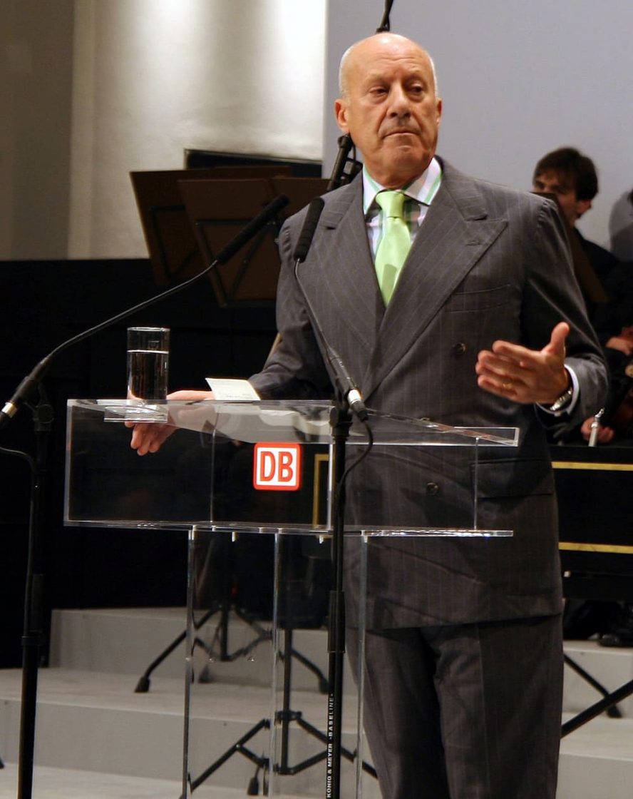 Norman Foster discursando em um evento oficial, usando terno cinza e gravata verde