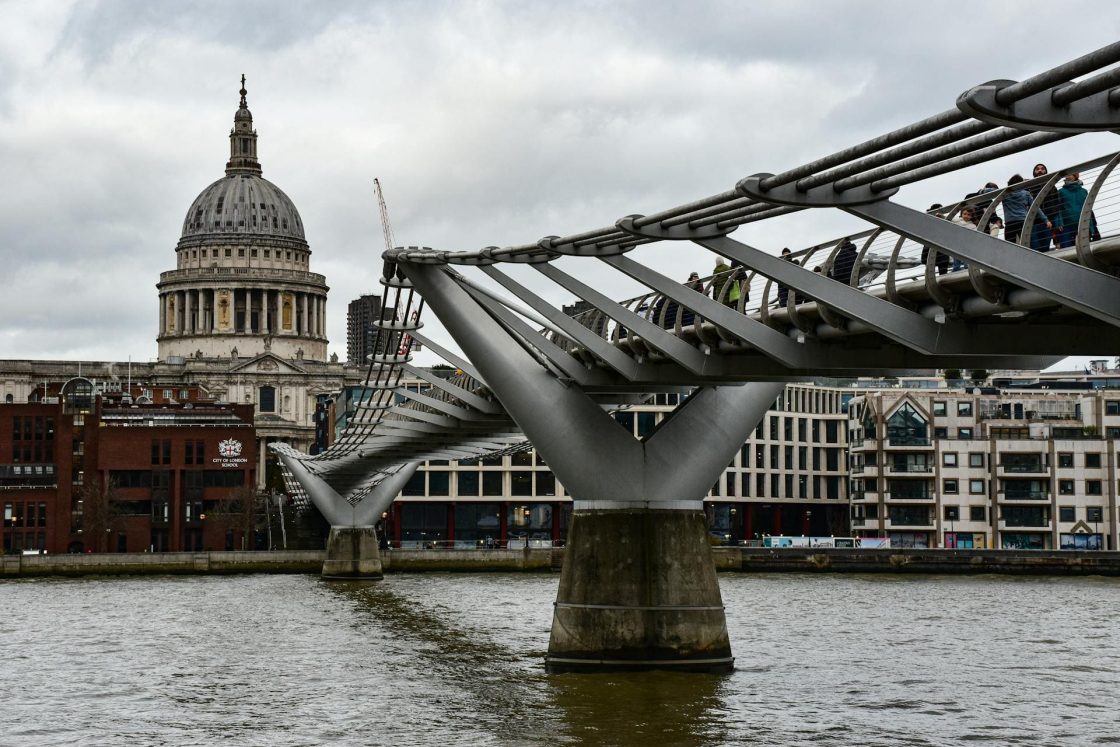 Vista lateral da Millennium Bridge atravessando o Rio Tâmisa, em Londres, com sua estrutura metálica sustentada por cabos e a cúpula da Catedral de St. Paul ao fundo