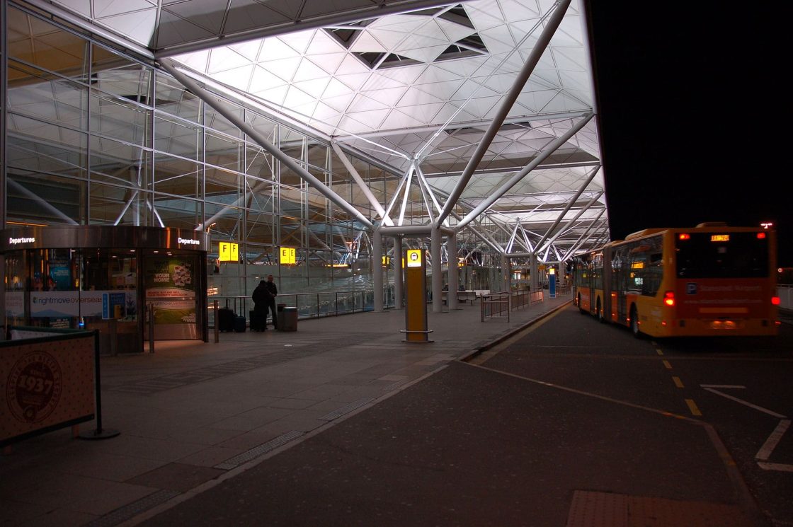 Terminal do Aeroporto de Stansted com cobertura geométrica em aço e vidro, vias de embarque de ônibus e áreas de circulação iluminadas