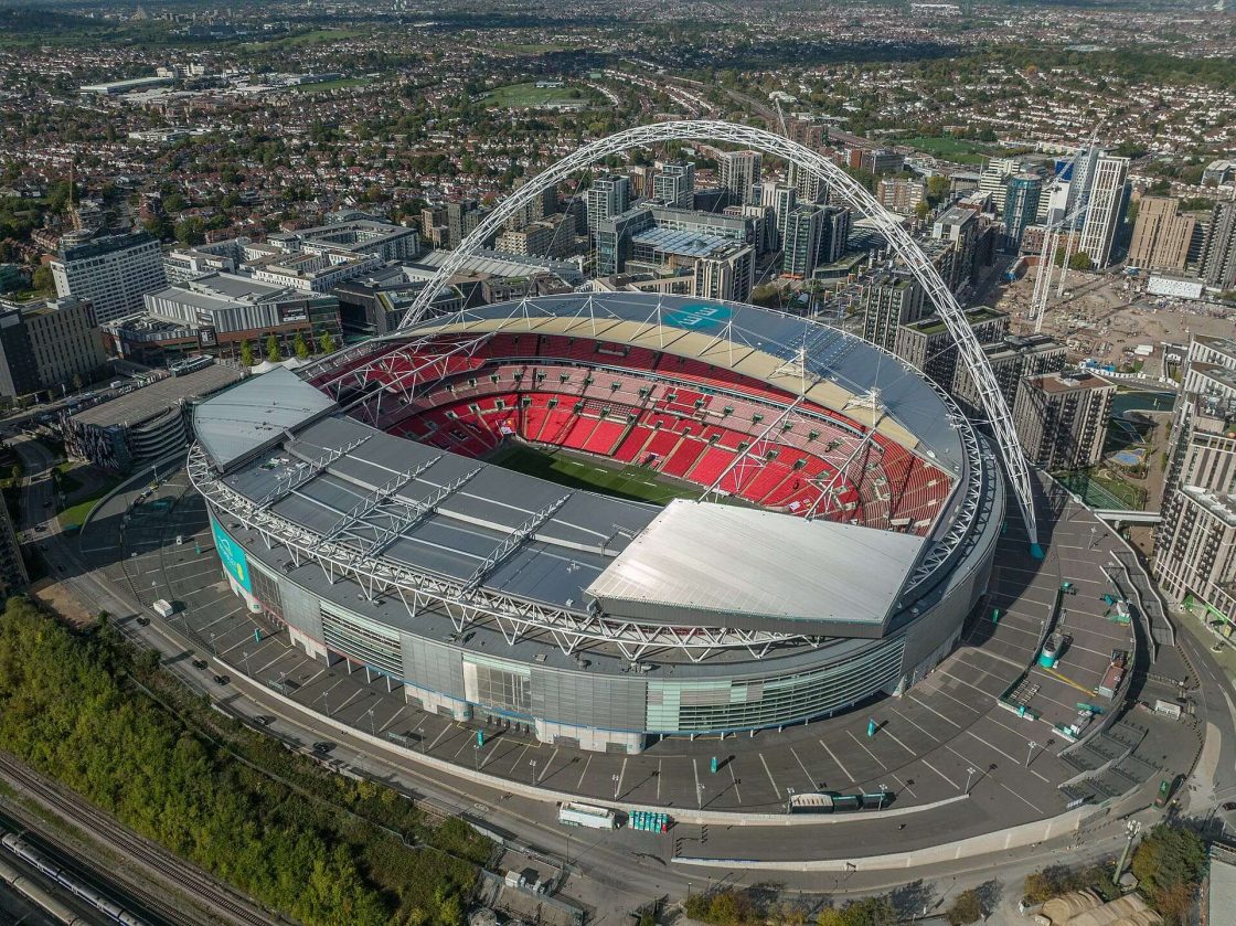 Vista aérea do Estádio de Wembley, em Londres, destacando o grande arco estrutural que atravessa a cobertura e a arena com arquibancadas vermelhas projetada com participação da Foster + Partners