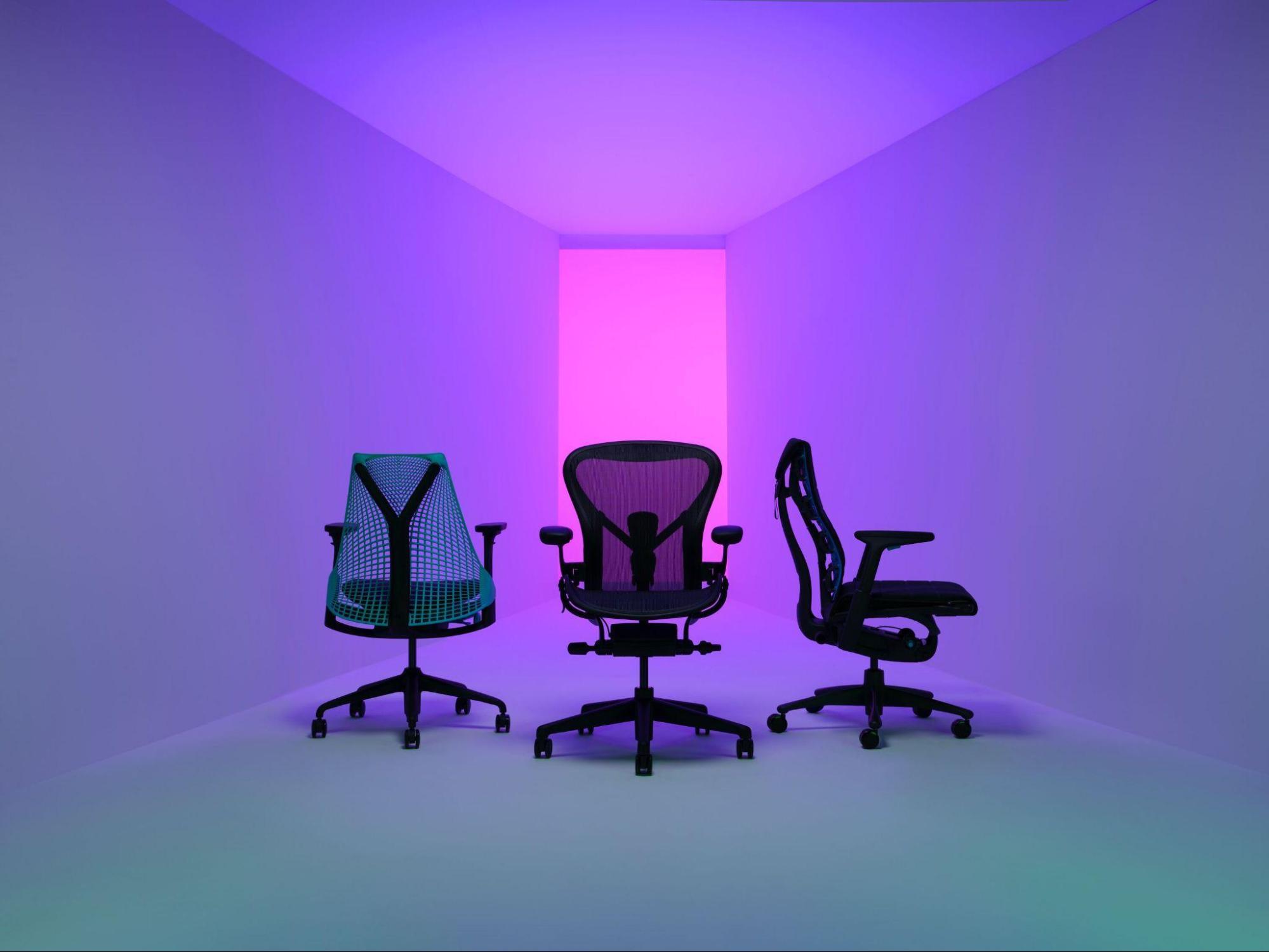 Fotografia de três cadeiras gamers pretas com elementos coloridos em espaço branco iluminado por uma luz violeta.