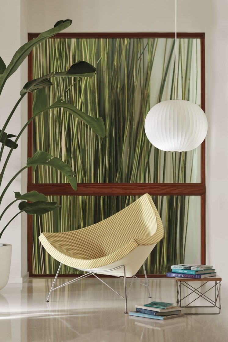  Fotografia de um ambiente interno com uma poltrona com estofado triangular modelo Coconut, da Herman Miller, um vaso de plantas, uma luminária branca, uma pilha de livros e outros objetos de decoração.