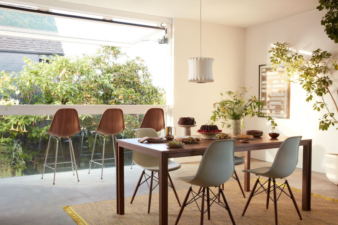 Fotografia de uma sala de jantar com mesa de madeira, cadeiras, alimentos, banquetas altas, luminária, quadro e vasos de plantas.