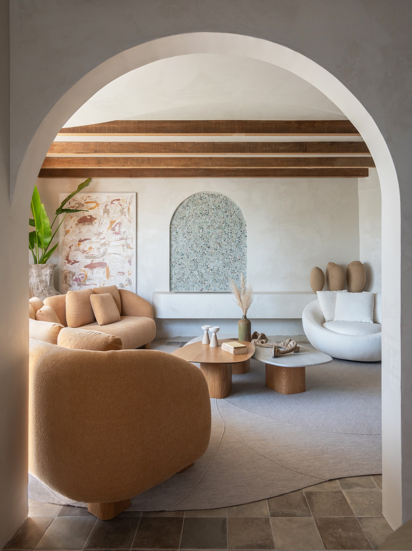 Sala de estar em estilo mediterrâneo emoldurada por arco. Sofás, poltrona e mesas em formas orgânicas. Quadros, vasos, esculturas e plantas