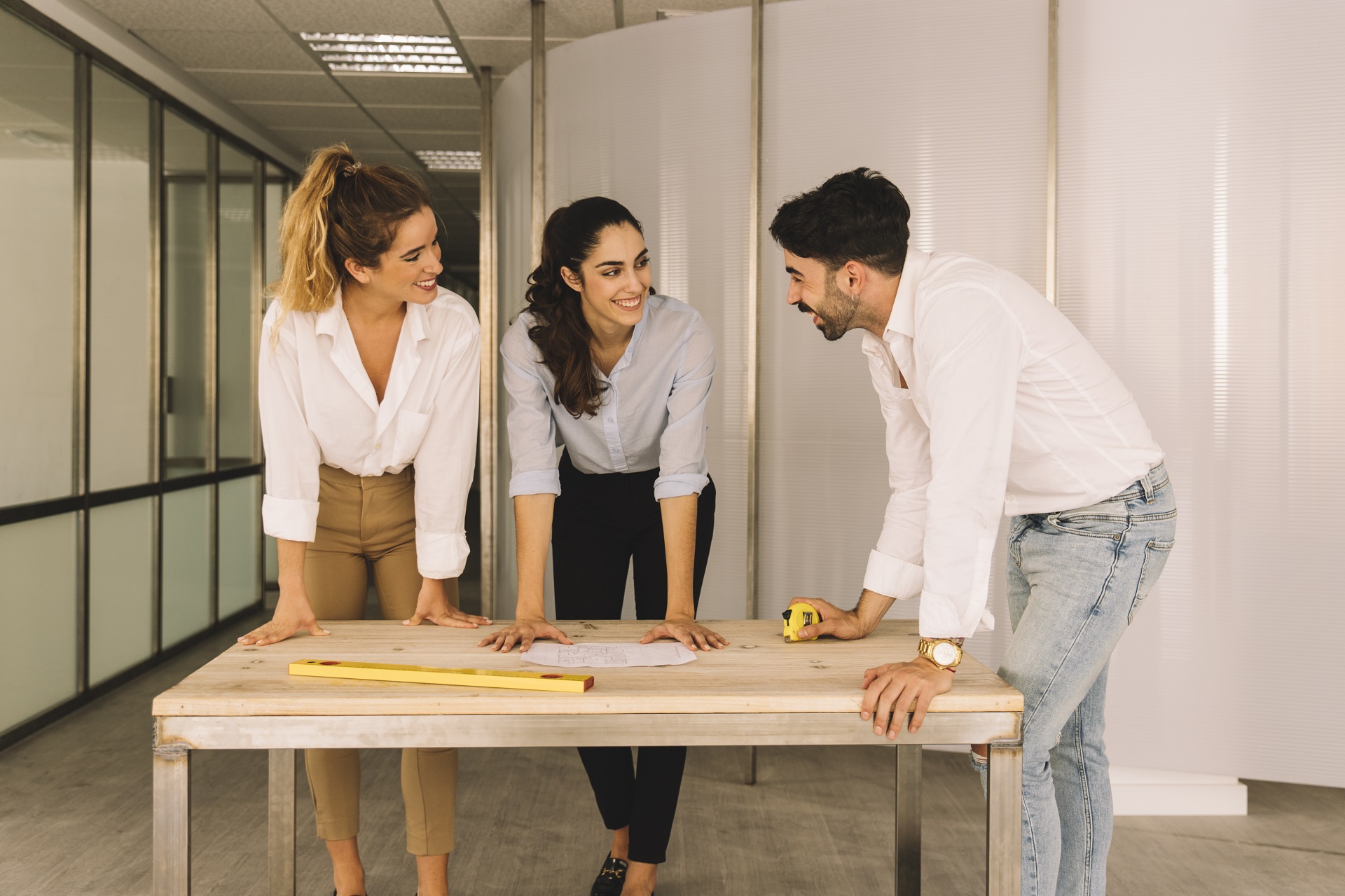 Duas mulheres e um homem, possivelmente arquitetos, discutindo um projeto sobre a mesa entre eles enquanto sorriem uns para os outros