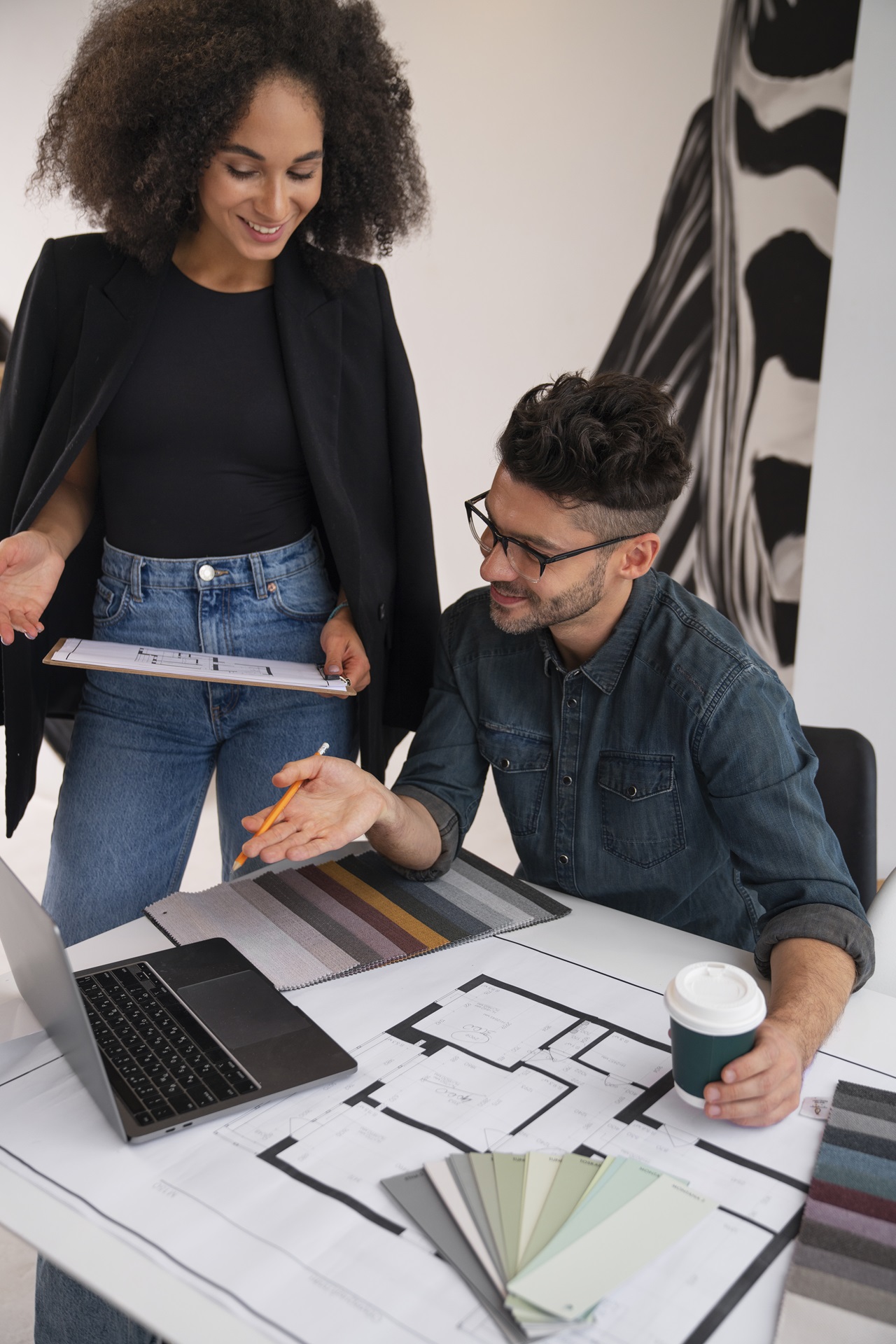 Um homem e uma mulher, ambos arquitetos, analisando um projeto. Há plantas em papel. Eles também usam um notebook e um tablet
