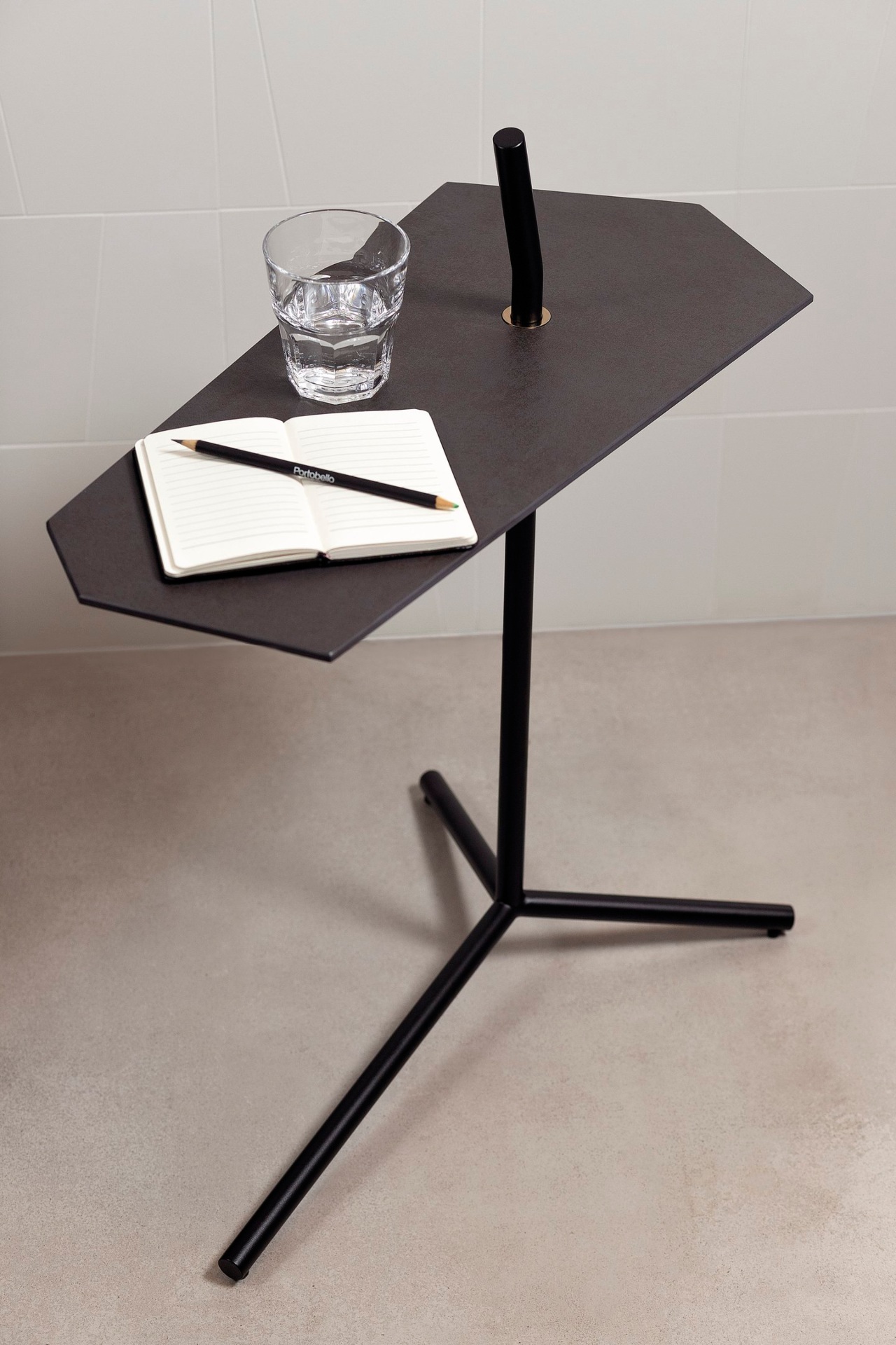 Mesa de canto com base de metal e tampo de porcelanato. Sobre ela há um caderno, um lápis e um copo d\'água