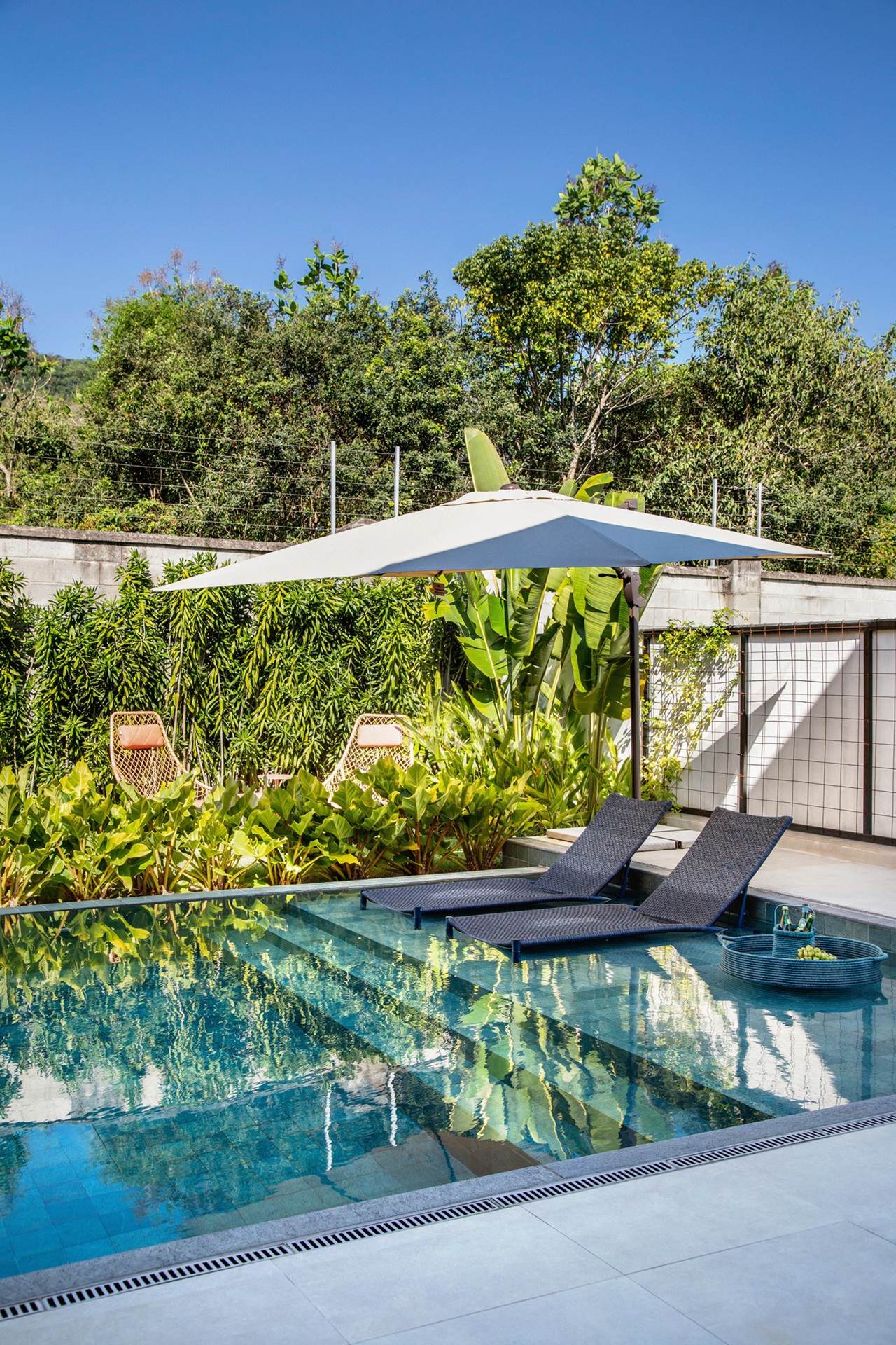 Piscina com deck molhado e duas espreguiçadeiras ao lado de um ombrelone com paisagimo abundante no entorno