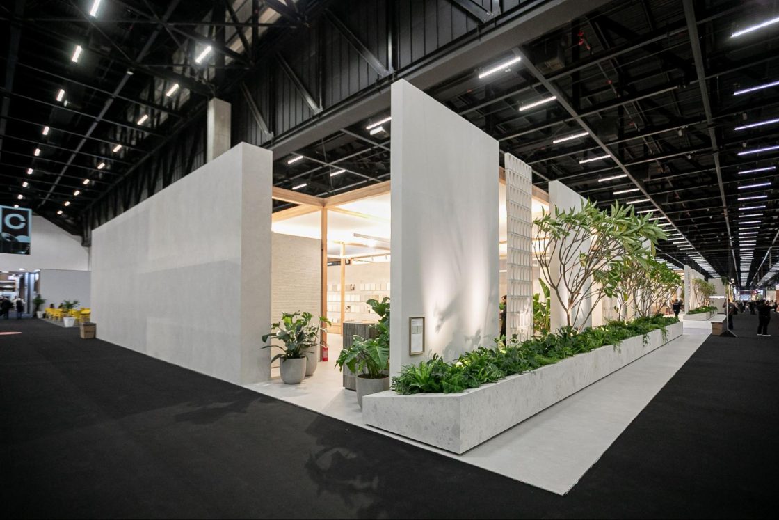 Fotografia com visão lateral de um estande em um espaço de exposições com paredes brancas e uma extensa floreira de plantas que circunda toda a lateral do ambiente.