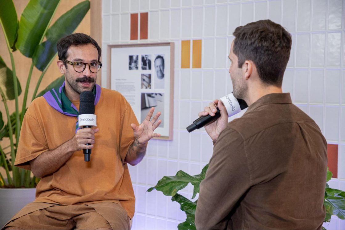 Fotografia de dois homens conversando sentados à frente de um painel de azulejos e vasos de plantas.