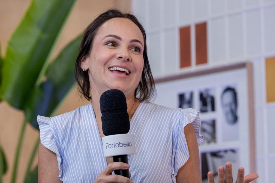 Fotografia de uma mulher de cabelos castanhos falando ao microofone à frente de um painel de azulejos e vasos de plantas.