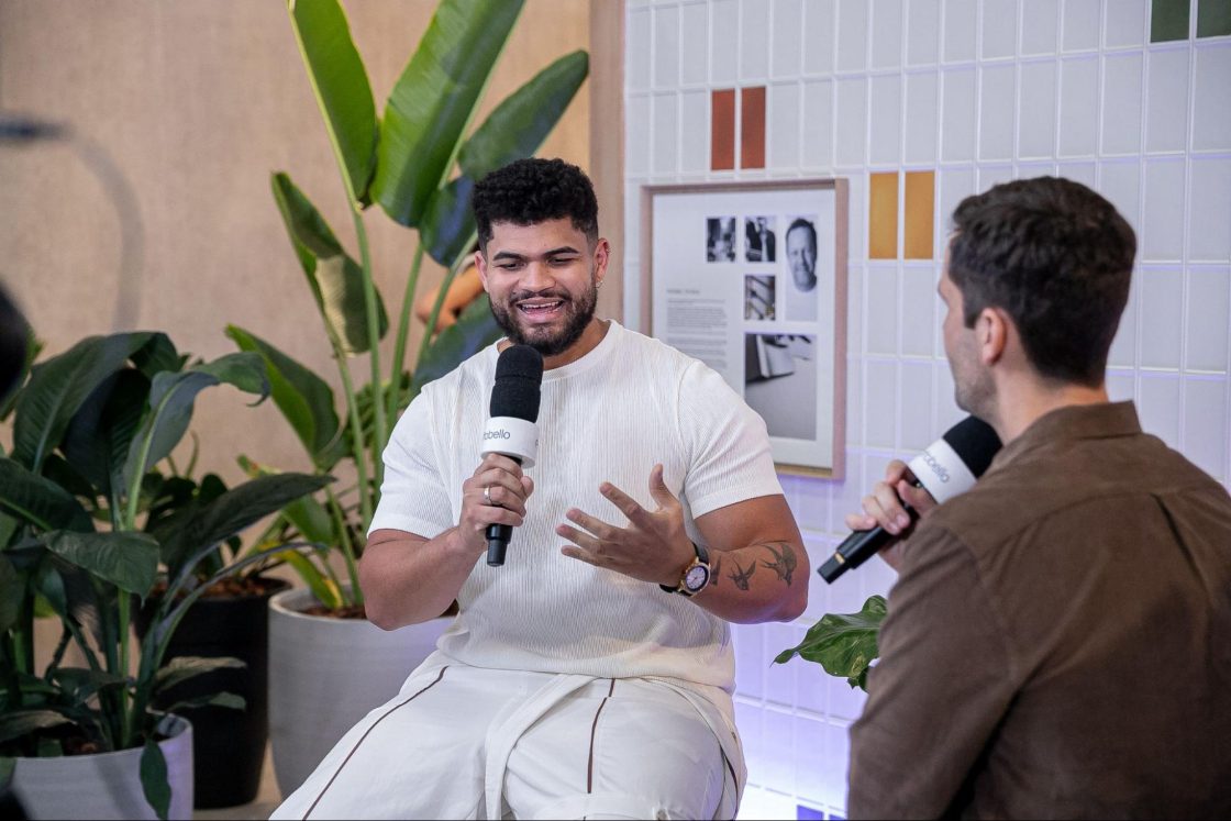 Fotografia de dois homens conversando sentados à frente de um painel de azulejos e vasos de plantas.