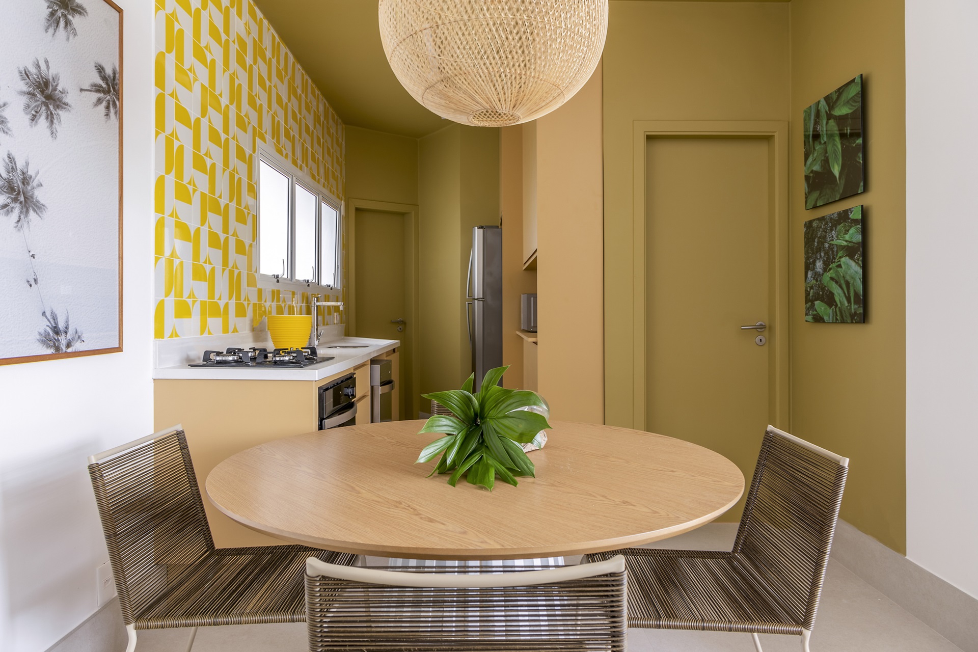 Sala de jantar integrada à cozinha com mesa redonda e quatro cadeiras. Na decoração, com predominância do amarelo, parede da esquerda tem revestimento decorado branco e amarelo