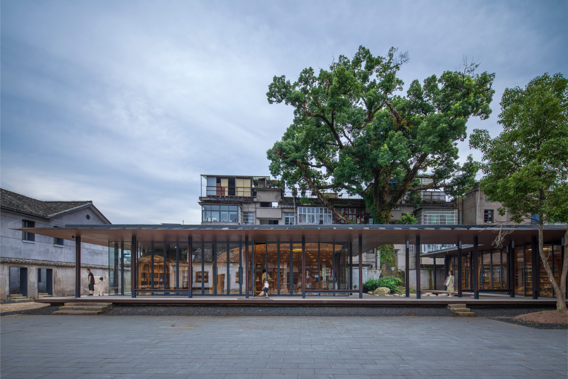 Centro Cultural de Songyang, edifício moderno de um pavimento, com estrutura envidraçada e linhas retas, situado em um ambiente urbano que mistura arquitetura tradicional e contemporânea. O prédio possui uma ampla cobertura plana que se estende além da estrutura
