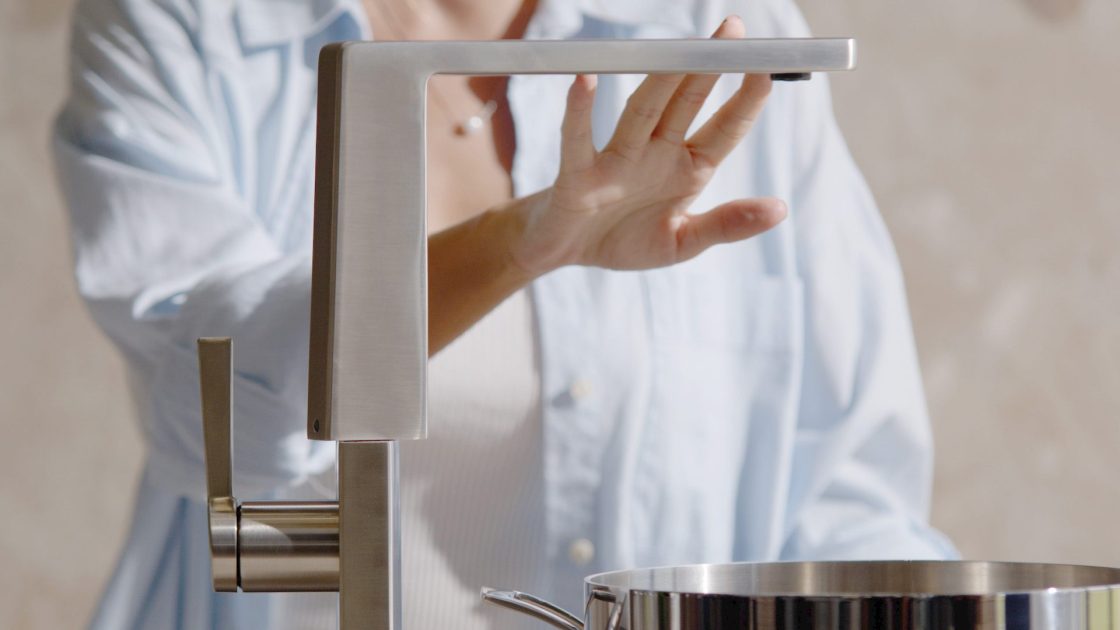 Mulher de camisa branca colocando a mão sobre torneira de bancada de cozinha para rotacionar a direção da bica