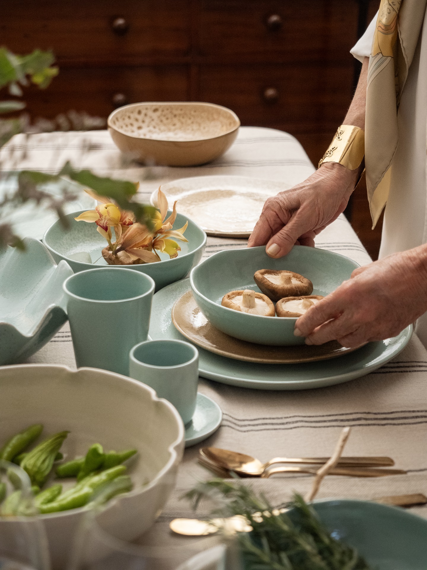 Mesa posta com louças azul-celeste. Mãos femininas aparecem segurando um prato fundo com cogumelos