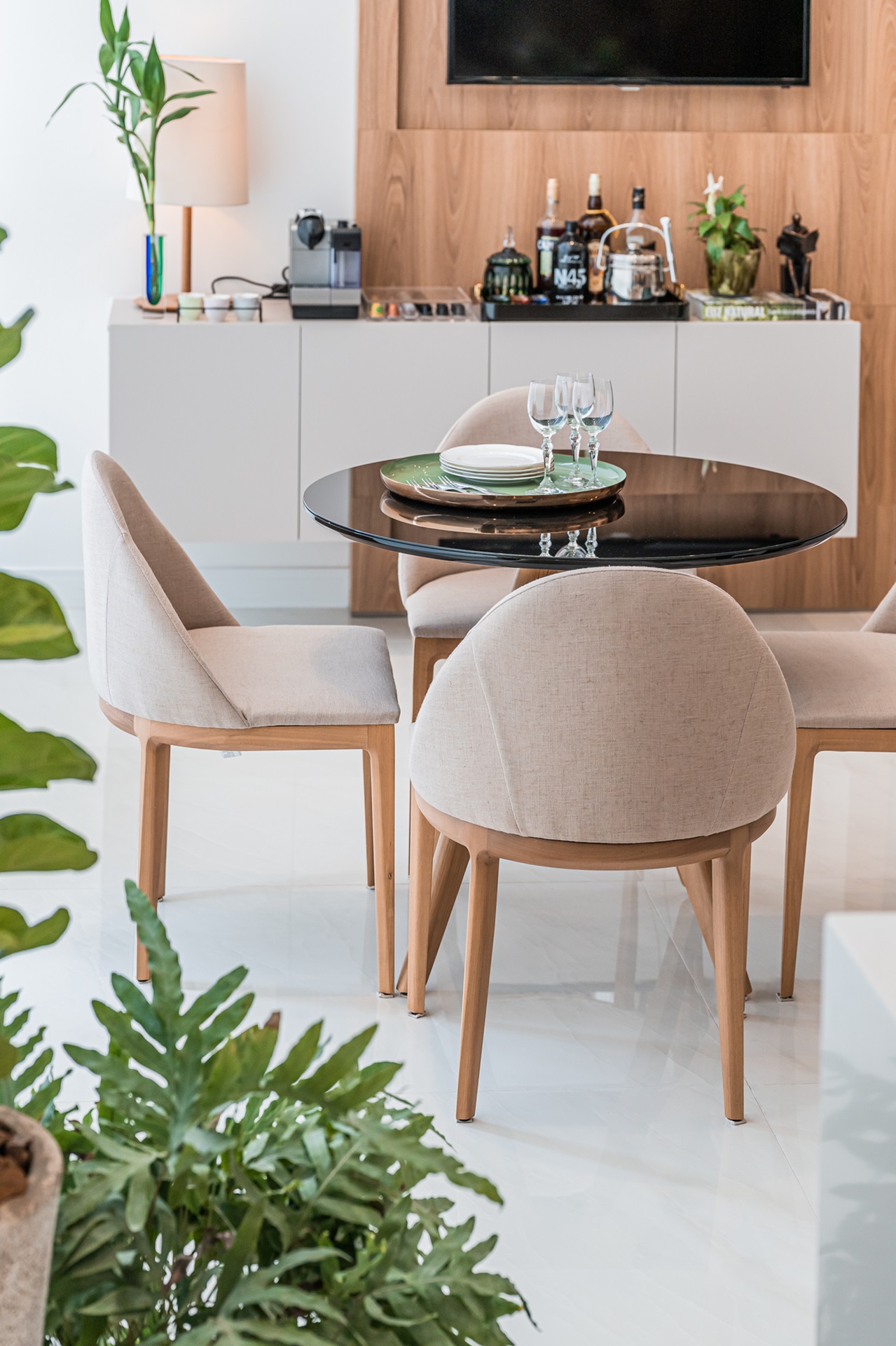 Sala de jantar com mesa redonda e quatro poltronas acolchoadas em tom de bege. Há um bar em buffet ao fundo