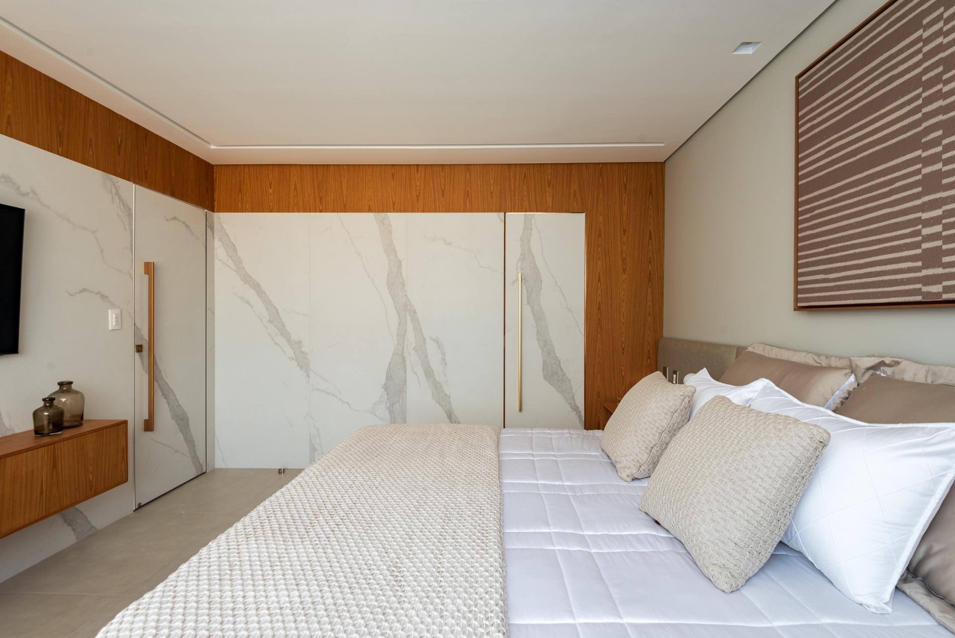 Cama king size em quarto. Ao fundo podemos ver um painel de madeira e porcelanato