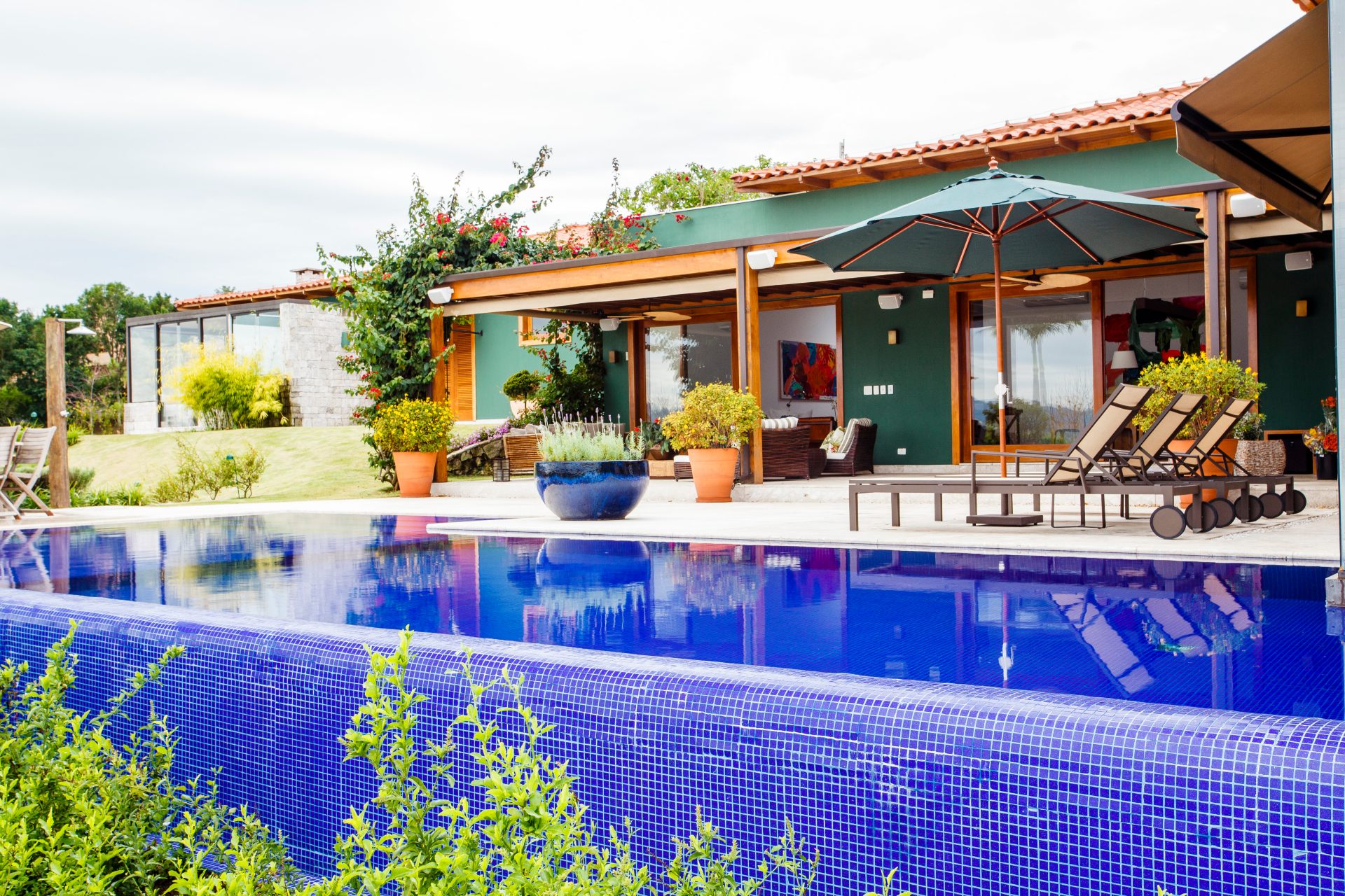 Em primeiro plano, há uma piscina de borda infinita revestida com pastilhas azuis. Ao fundo, a casa possui um design rústico e acolhedor, com paredes pintadas em um tom de verde-escuro, estrutura de madeira e um telhado de telhas de barro