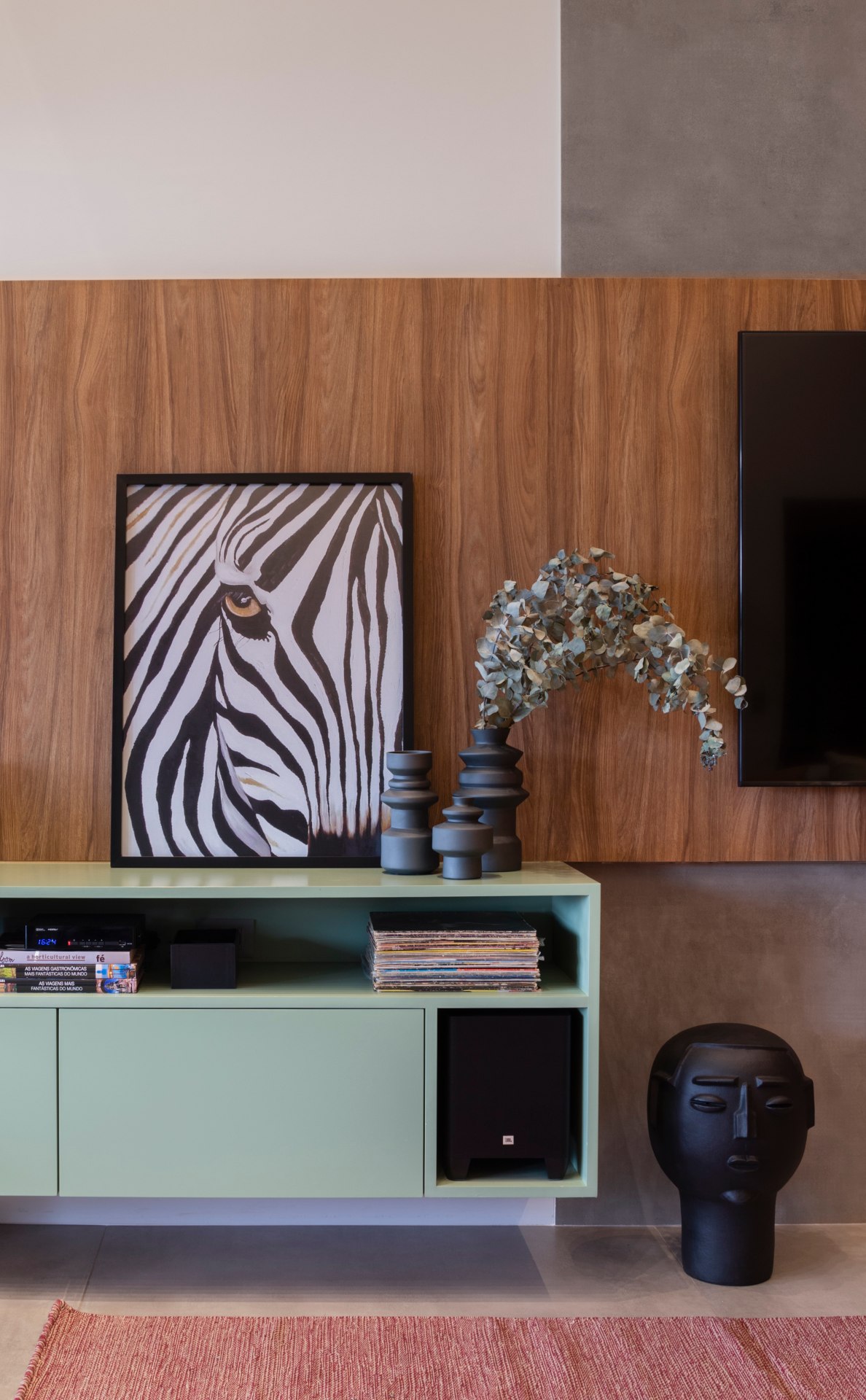 Parte de uma sala de estar em que é possível ver na parede ao fundo parte de uma televisão fixada num painel de madeira, um rack verde claro com um quadro que remete a uma zebra e um conjunto de vasos sobre ele. No chão, ao lado do rack, uma escultura de cerâmica com formato de cabeça, feita por Mestre Neguinha