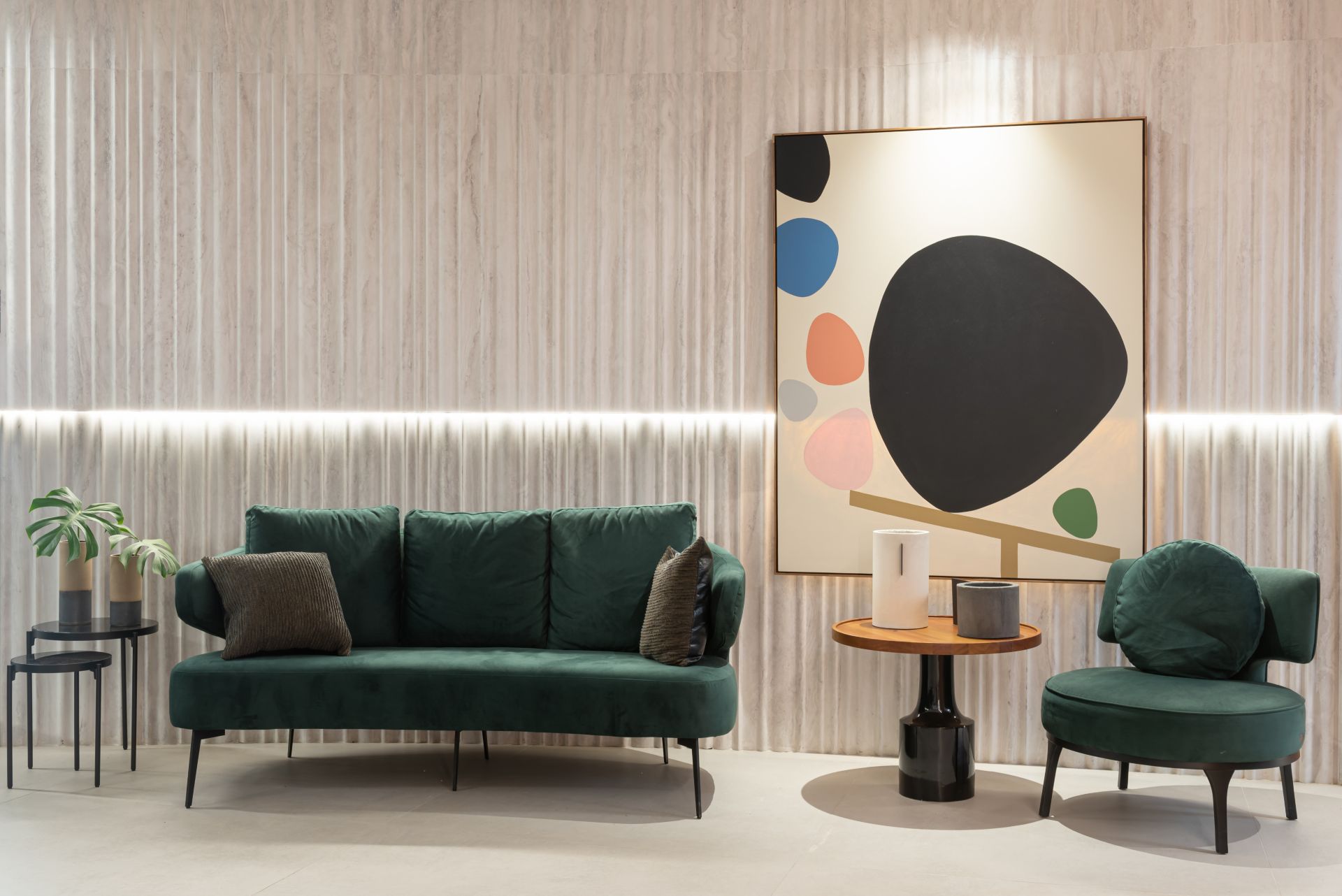 Ambiente de uma sala de estar, com um sofá de três lugares e uma poltrona verdes, separados por uma mesinha de canto. Na parede, no canto direito, há um quadro de fundo branco com formas orgânicas arredondadas de diferentes cores