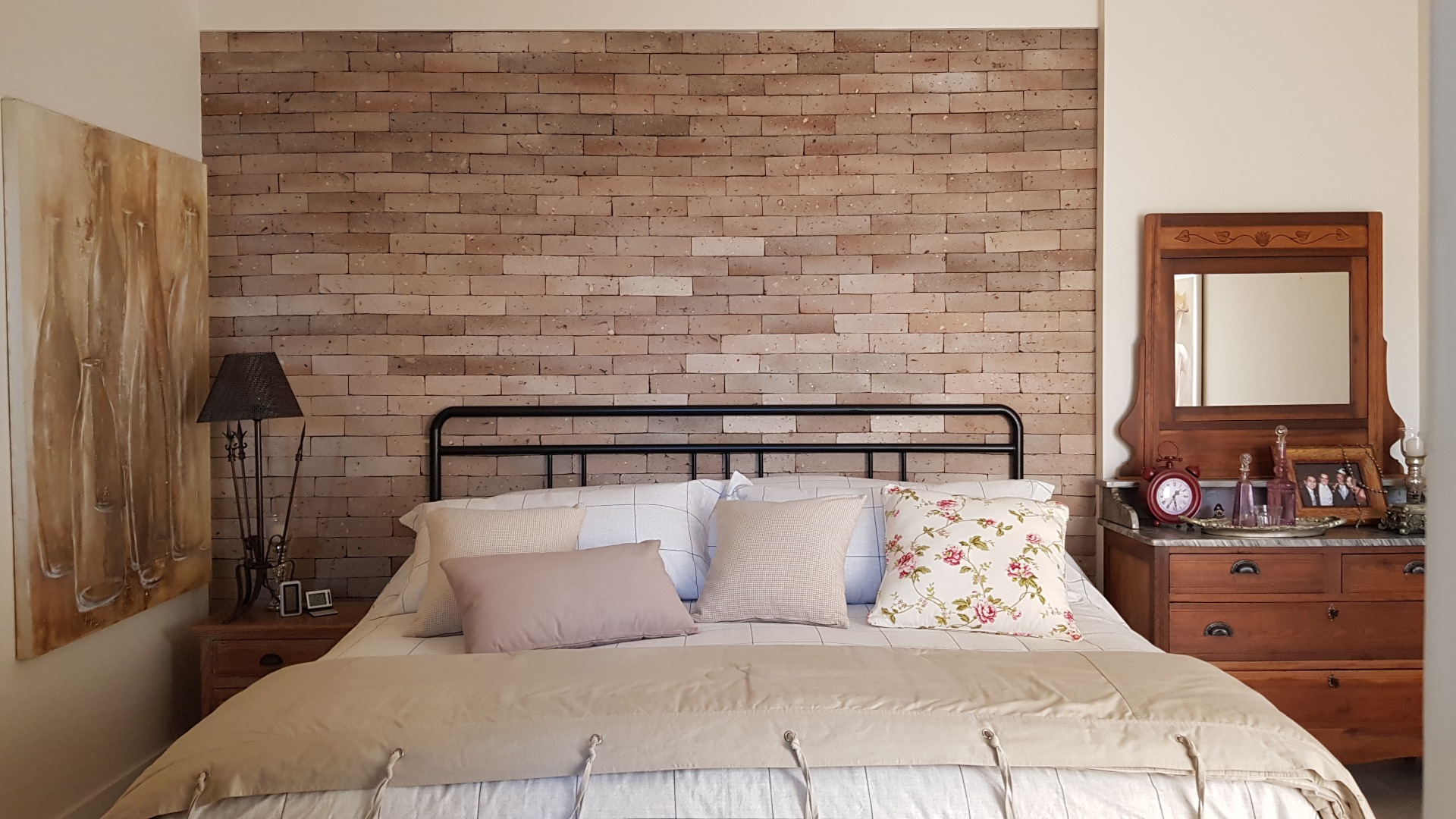 Quarto com móveis antigos de madeira, parede do fundo de tijolinhos e uma cama de casal decorada com jogo de cama e almofadas