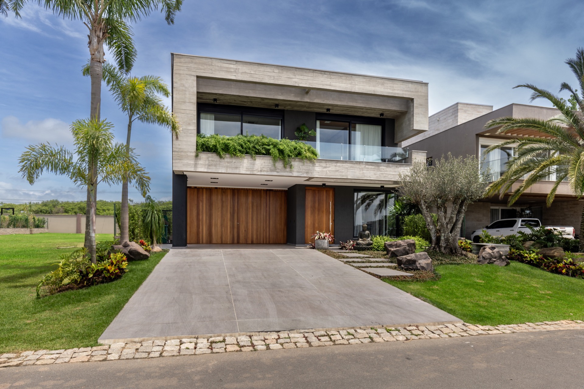 Vista da fachada de casa grande de dois andares em condomínio. Com design moderno, possui uma rampa para veículos na entrada. No primeiro andar, a fachada conta com madeira e vidro. No segundo andar, há uma ampla varanda com portas e guarda-corpo em vidro