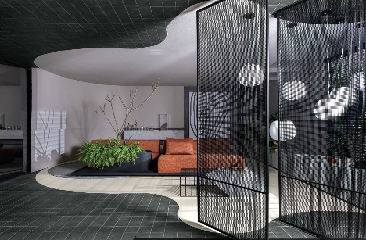 Ambiente demarcado por supercífices brancas no teto, no piso e nas paredes. Conta com um sofá marrom, luminárias modernas penduradas, quadro ao fundo e plantas que compõem a decoração