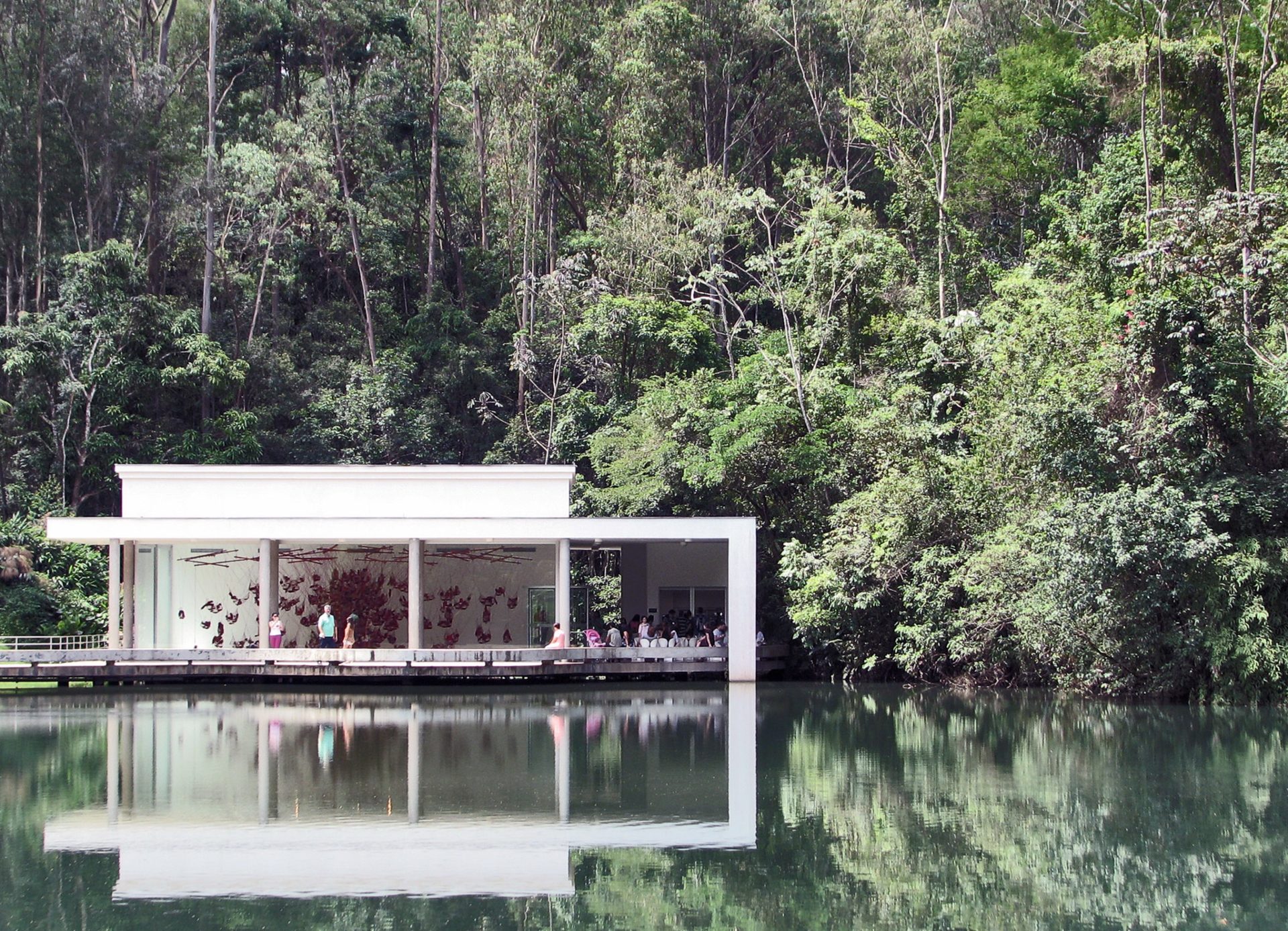 Vista para uma das galerias do Instituto Inhotim, refletida no lago a frente e com árvores ao fundo. É possível enxergar a obra exposta na parte interna, com diversas peças vermelhas penduradas, e alguns visitantes