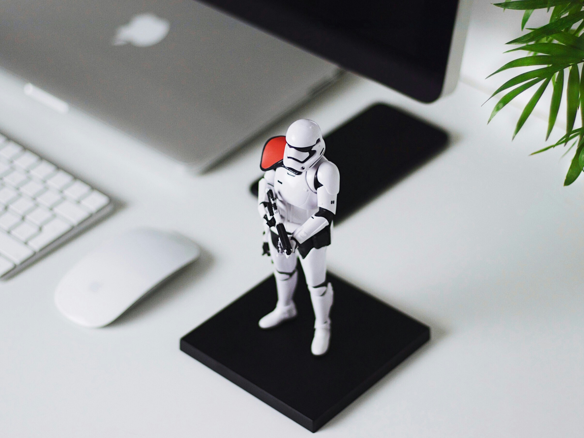 Mesa branca com um notebook da Apple, teclado, mouse e um boneco Stormtrooper em destaque sobre uma base preta. Há também uma planta no canto
