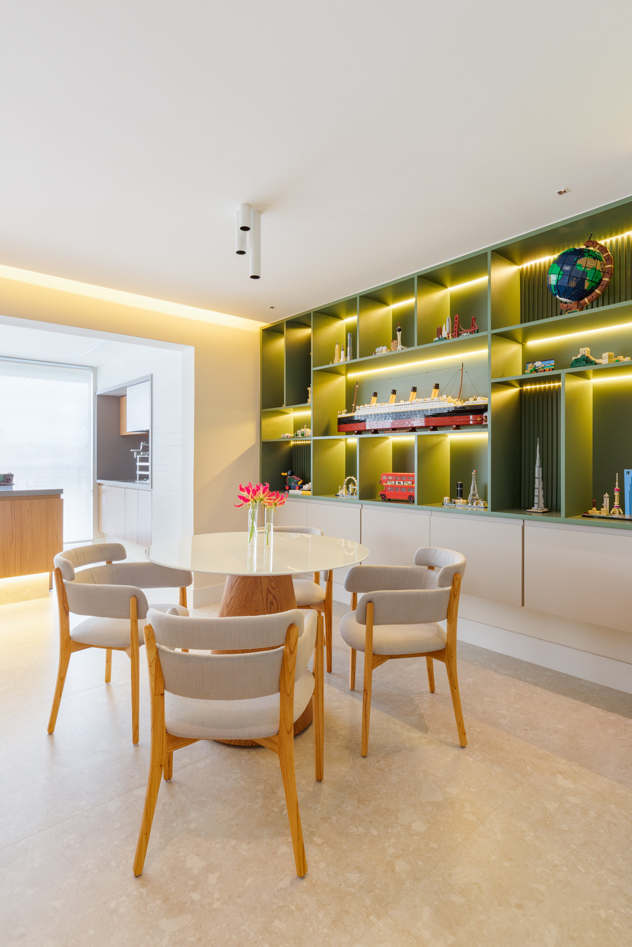 Sala de jantar moderna e iluminada, com uma mesa redonda branca e quatro cadeiras de madeira com estofado claro. Ao fundo, há uma estante verde com nichos iluminados, decorada com modelos de Lego de monumentos e um navio, além de outros objetos decorativos