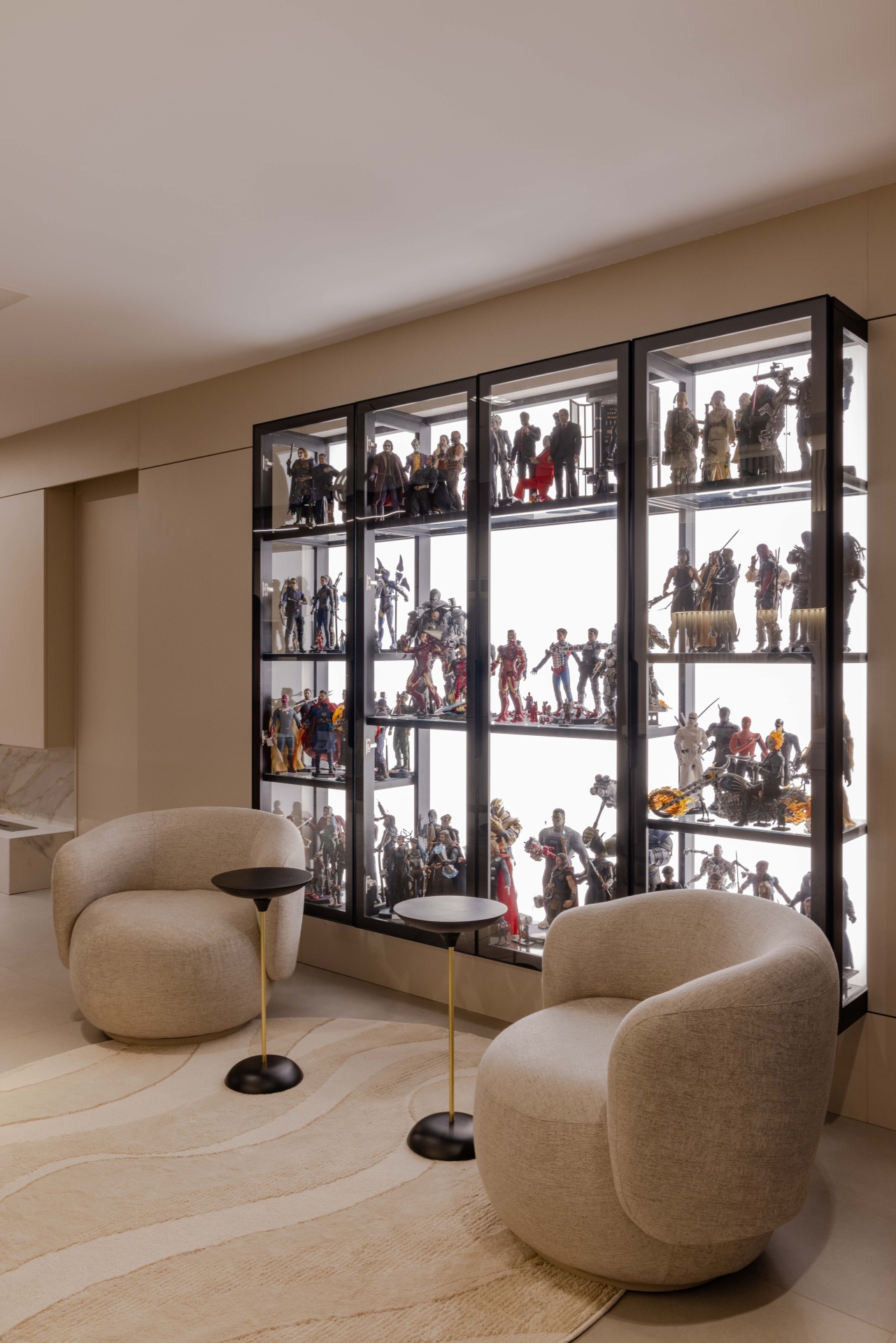 Uma estante com portas de vidro exibe action figures de diversos universos fictícios. À sua frente, poltronas na cor creme