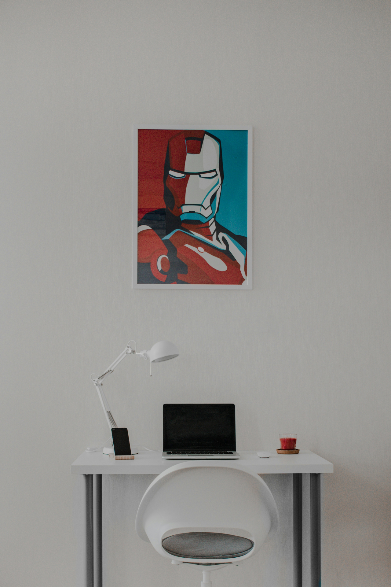 Escritório minimalista com uma mesa branca, um notebook, uma luminária, um celular e um copo. Acima da mesa, há um pôster estilizado do Homem de Ferro na parede