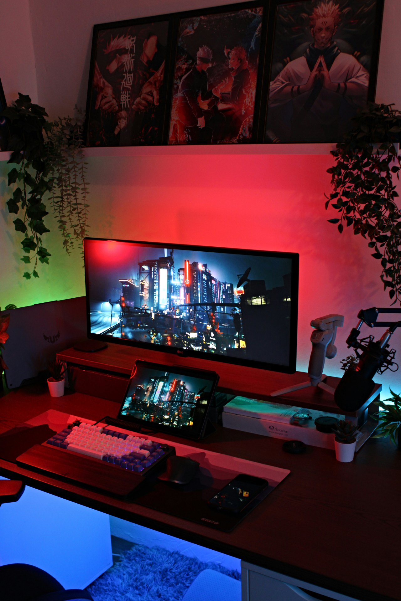Uma mesa com computador e tablet. Atrás do monitor há uma luz LED vermelha, enquanto do lado esquerdo há uma luz led verde. Acima da mesa, existe uma prateleira com alguns quadros 