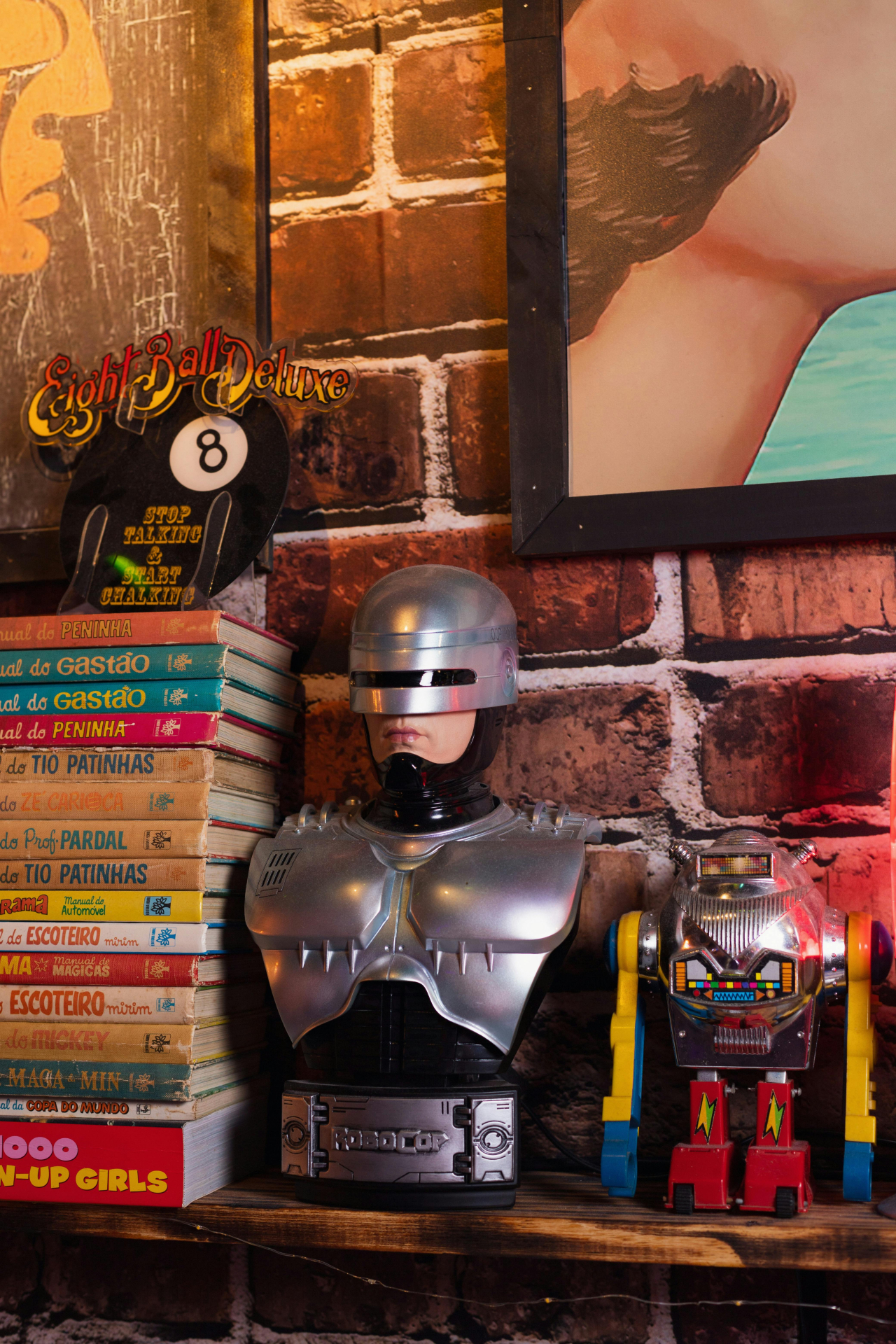 O busto do personagem Robocop está em destaque. No lado esquerdo, há alguns livros, enquanto no lado direito existe a presença de um robô colorido. Atrás há uma parede de tijolos com dois quadros