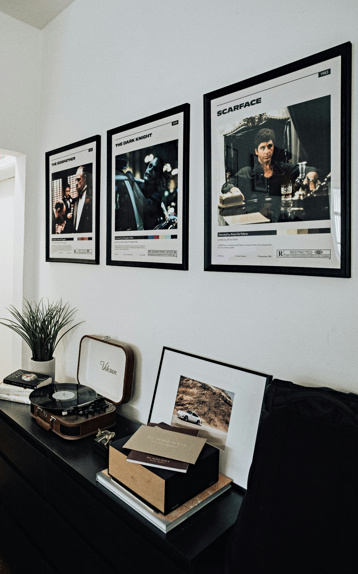 Parede branca com quadros que contêm pôsteres de filmes clássicos. Abaixo dos quadros há um aparador preto que sustenta uma vitrola, vaso com planta e mais um quadro 