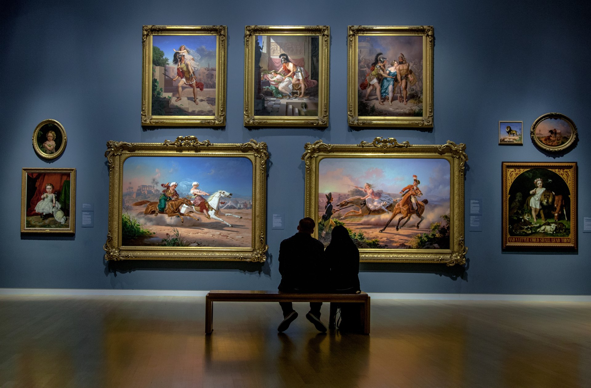 Casal sentado sobre um banco em um museu admira quadros expostos na parede ao fundo