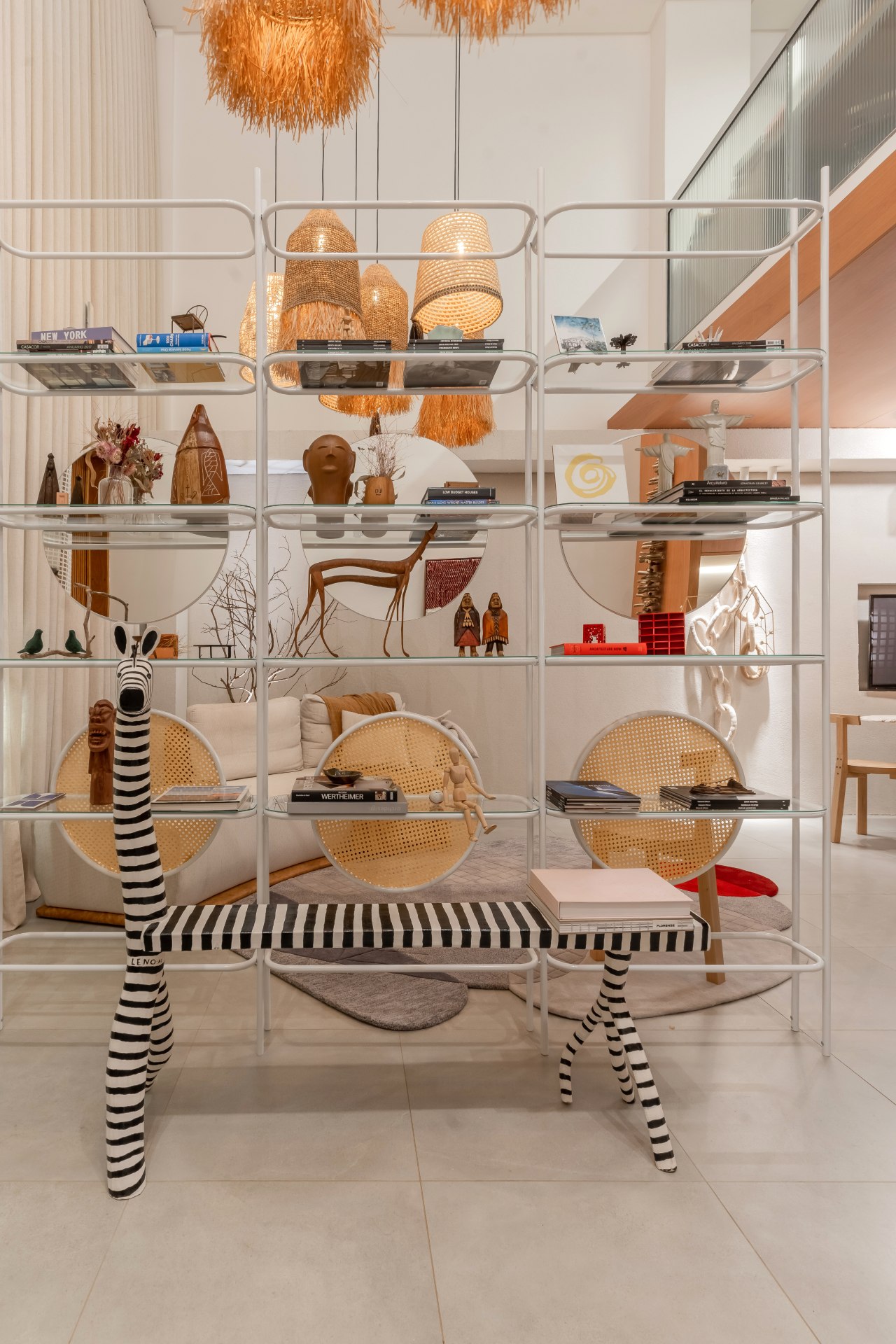 Estante de metal com diversos objetos decorativos artesanais e livros, além do papel decorativo, funciona como uma espécie de divisória na sala de estar. Em frente à estante, destaca-se um banco de madeira artesanal que reproduz uma zebra
