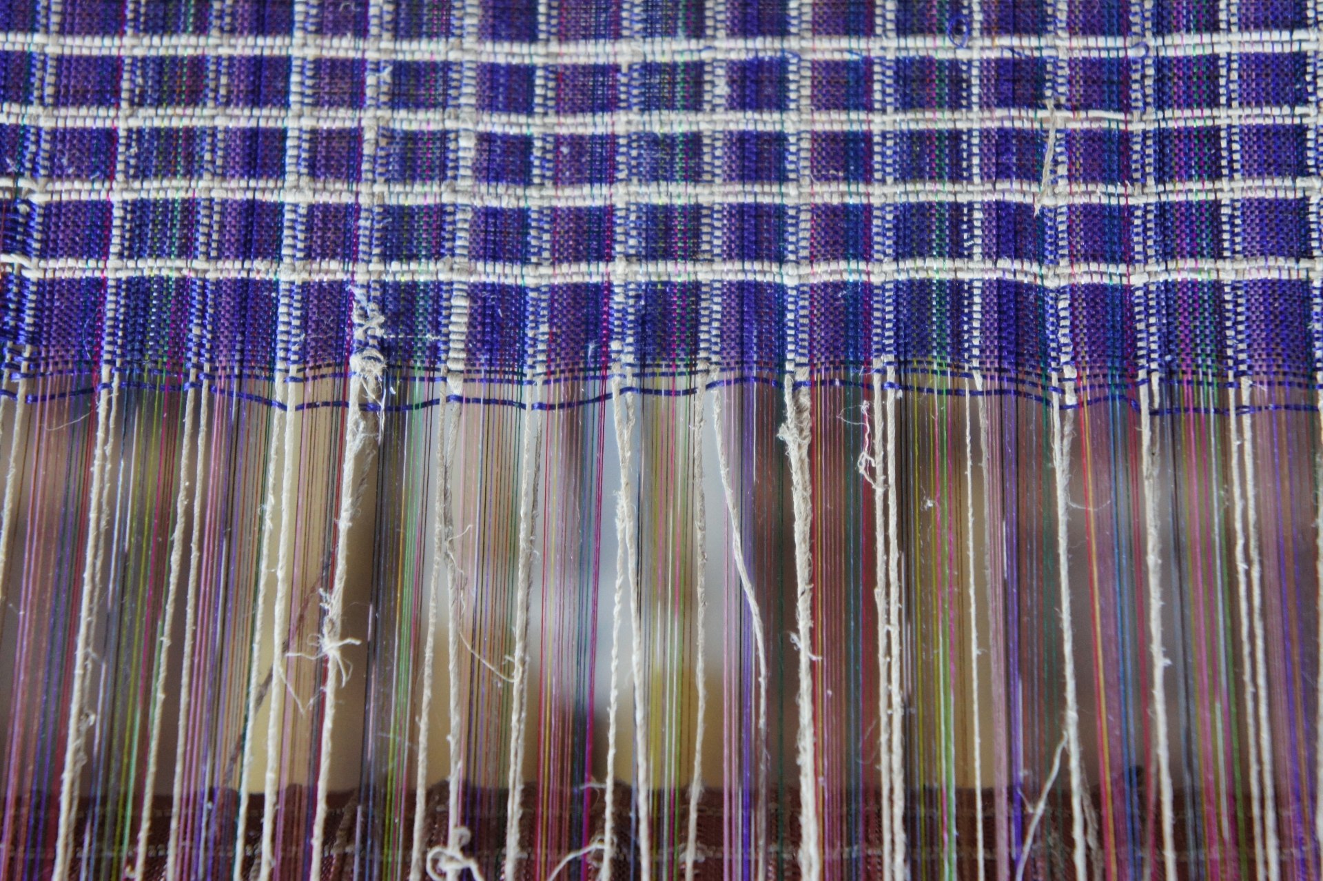 Fios sendo entrelaçados manualmente por meio de tear. Em metade da foto, os fios estão esticados; na outra metade, estão dando origem a um tecido quadriculado azul
