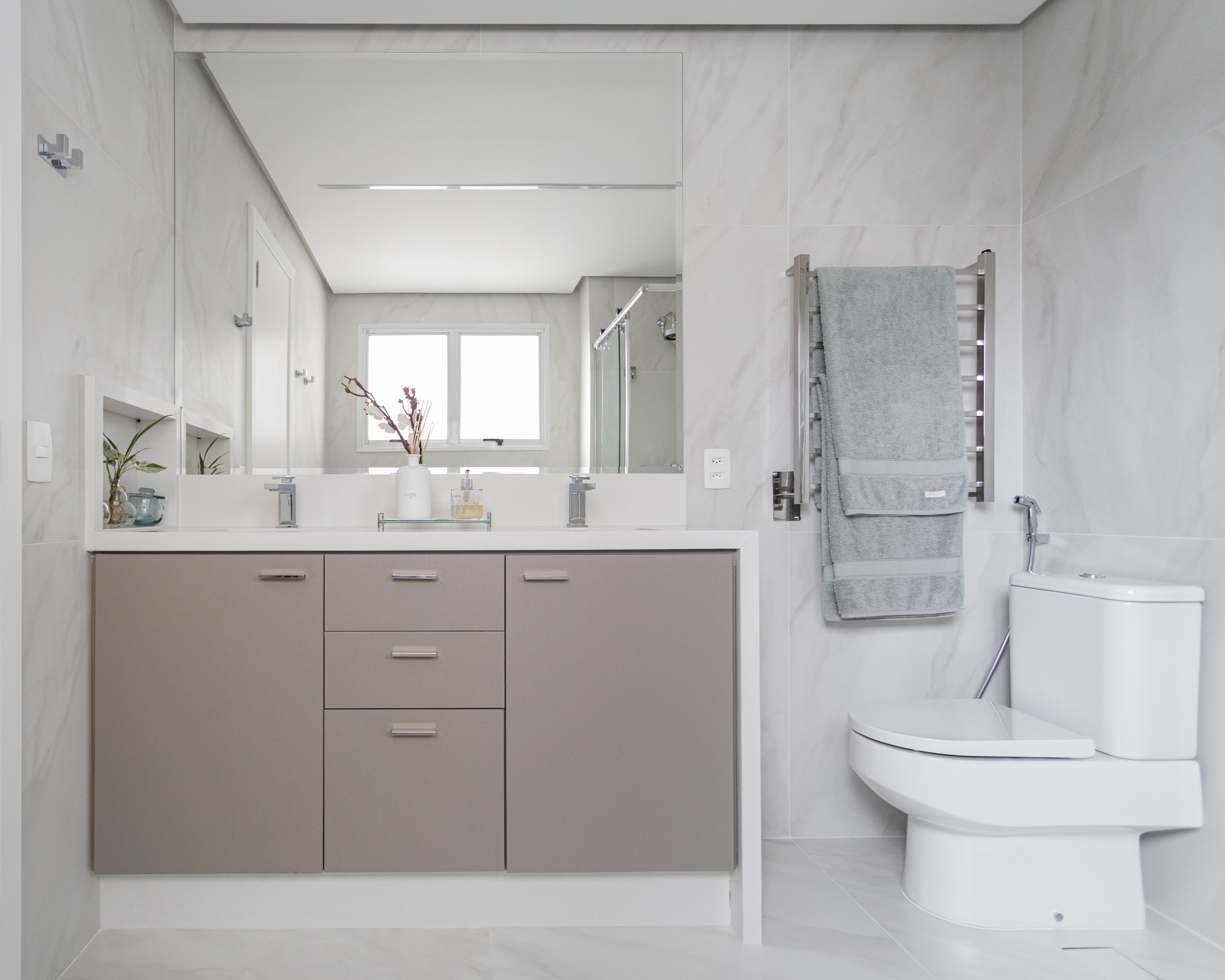 O banheiro apresenta um vaso sanitário branco e uma bancada, com detalhes elegantes em seu design. Em um close, é possível ver uma toalha cuidadosamente dobrada, um armário e uma planta em um vaso de vidro, enquanto uma porta de vidro para o chuveiro e uma janela complementam a atmosfera do ambiente