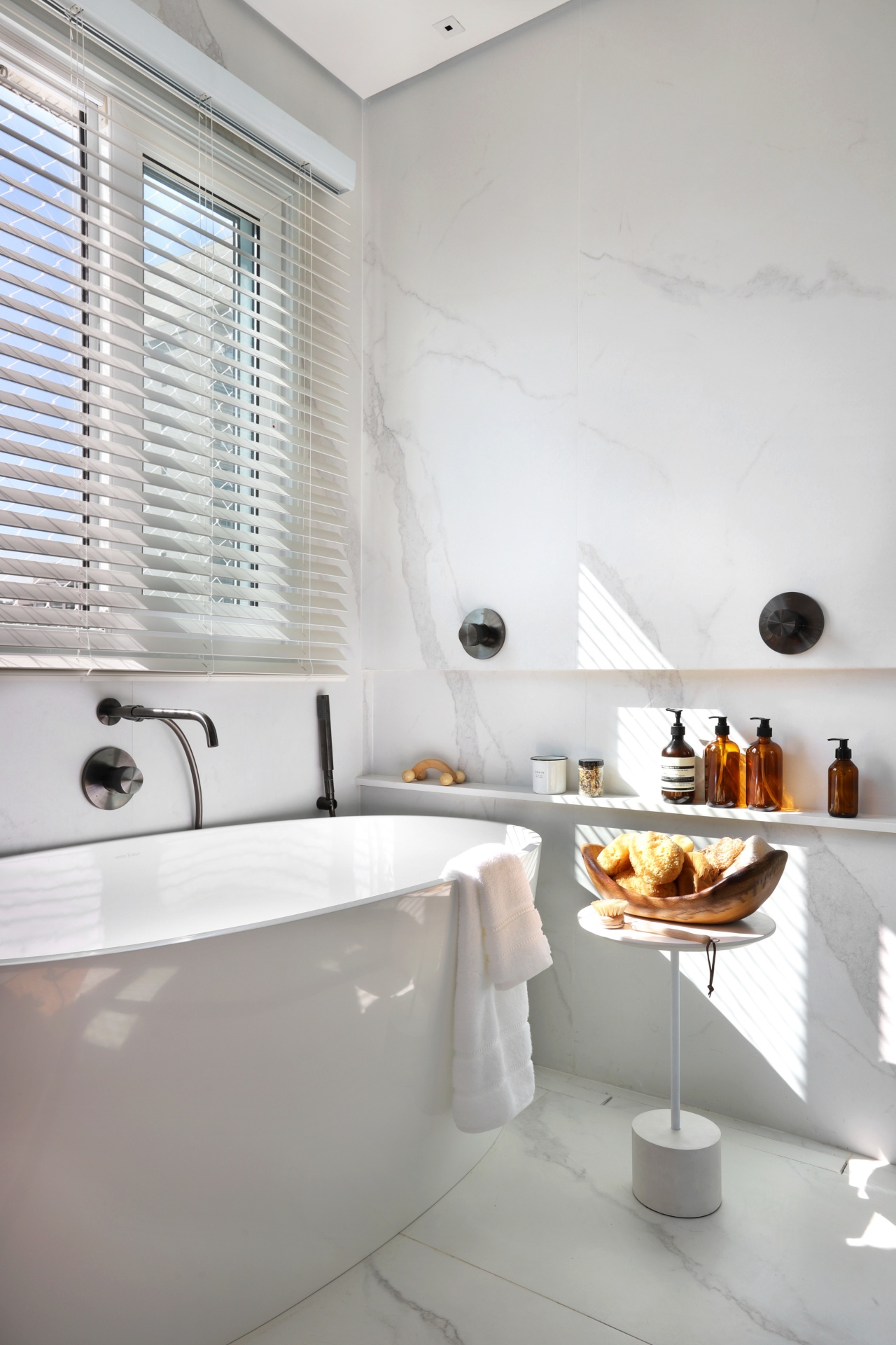 Banheiro com piso e paredes em porcelenato que reproduz mármore. Podemos ver uma banheira de imersão, uma mesinha e um nicho com produtos de higiene e beleza