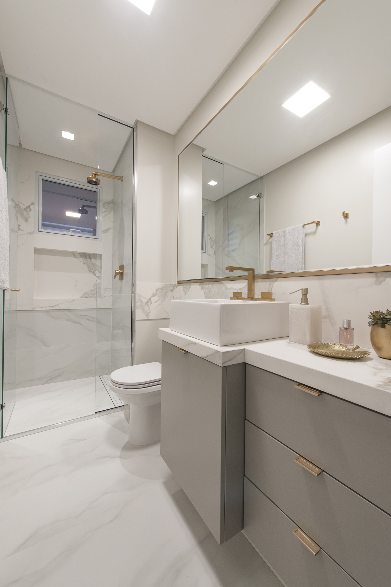 O banheiro conta com um chuveiro e uma bancada elegante, com a luz suave. Detalhes incluem um vaso sanitário, uma planta e uma torneira dourada 