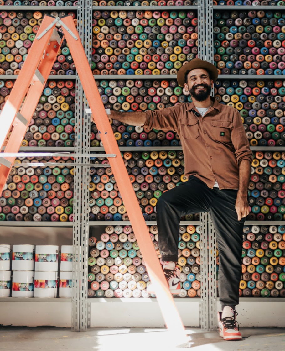 Eduardo Kobra, sorridente, encostado em uma escada laranja, posa em frente a prateleiras cheias de latas de spray coloridas