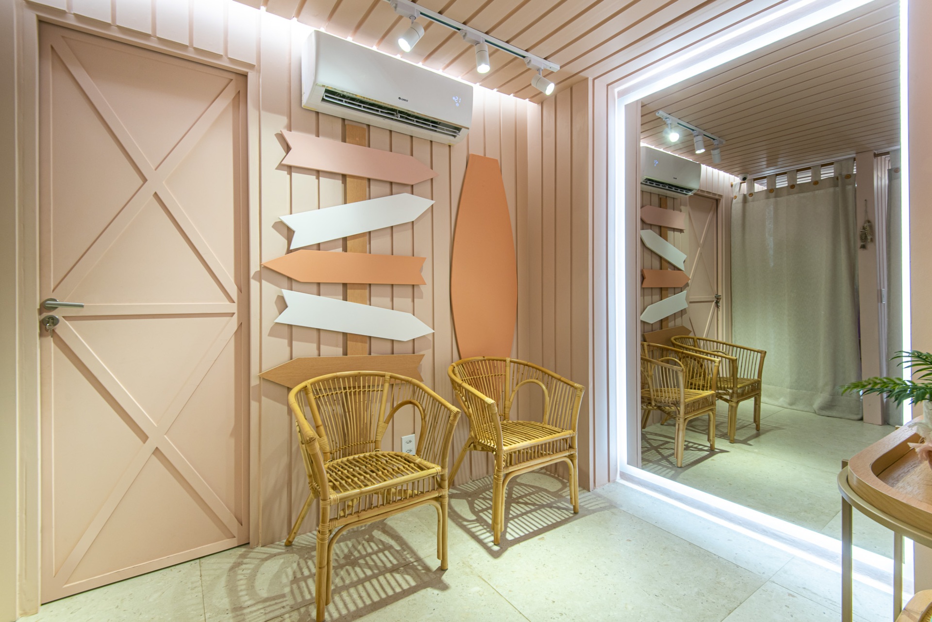 Cadeiras de vime e elementos simples, como um espelho, ar-condicionado branco e parede rosa. O detalhe das setas na parede adiciona um toque de modernidade e dinamismo ao espaço.