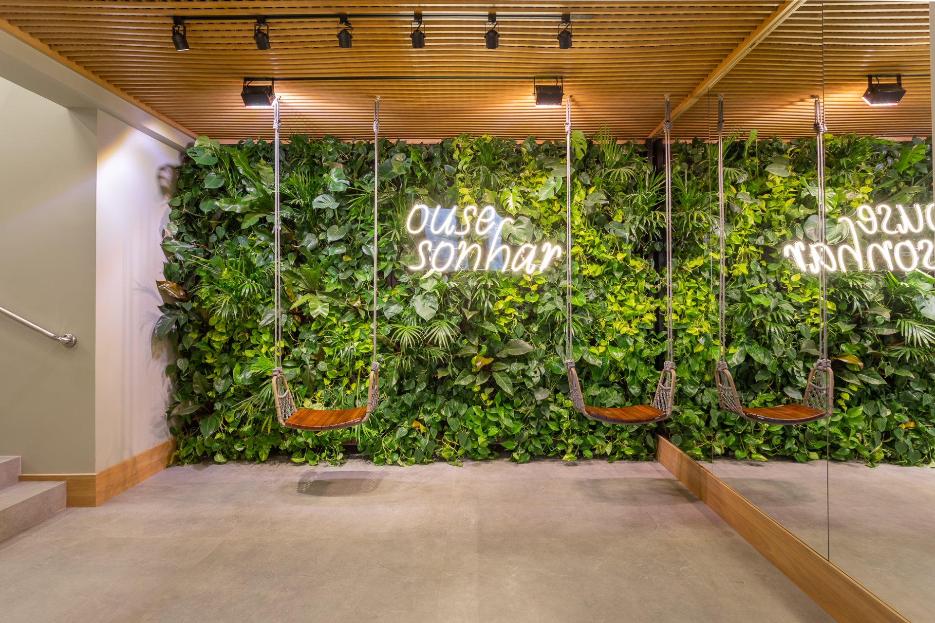 As plantas verdes, os balanços suspensas e a placa de neon criam uma atmosfera vibrante e relaxante. O piso de concreto e os detalhes como o balanço de madeira contrastam com os elementos orgânicos