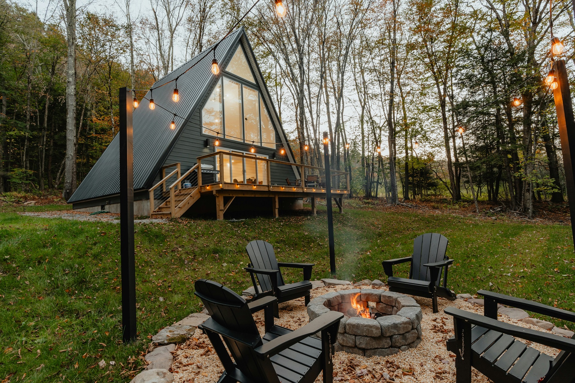 Em frente a uma charmosa cabana triangular no meio da floresta, um fire pit de pedras é cercado por cadeiras de madeira e iluminado por cordões de luz