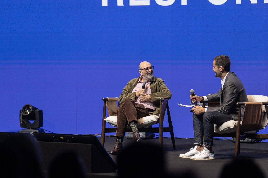 Isay Weinfeld e Pedro Andrade sentados em poltrona, segurando microfones, em palco do Archtrends Summit