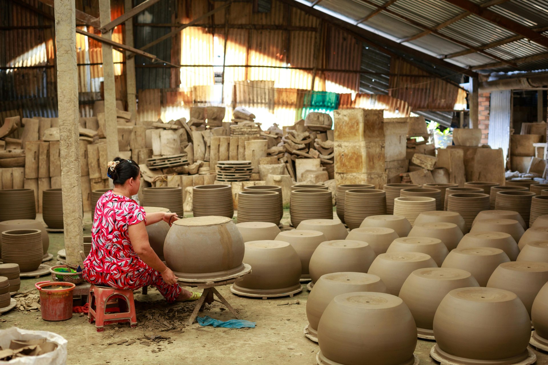 Mulher sentada em um pequeno banco, produzindo vaso artesanal de cerâmica em um galpão cheio de outros vasos grandes em processo de secagem